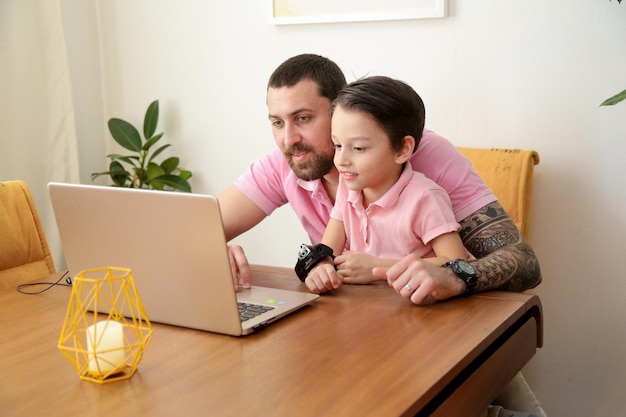 Giovane padre felice in polo rosa che lavora al computer portatile mentre è seduto al tavolo con il suo piccolo figlio famiglia felice che lavora a casa concetto