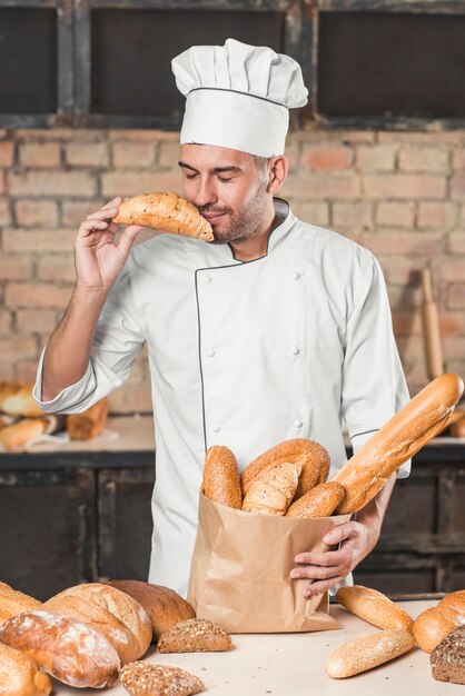 Giovane panettiere maschio sorridente che sente l&#39;odore del croissant fresco con i pani