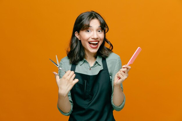 Giovane parrucchiere donna che indossa grembiule tenendo pettine per capelli e forbici guardando la fotocamera felice e positivo sorridente allegramente in piedi su sfondo arancione