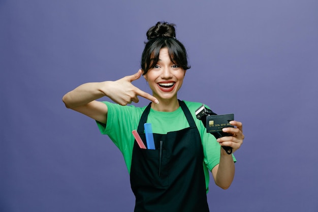 Giovane parrucchiere donna che indossa un grembiule con in mano rasoio elettrico e carta di credito puntata con il dito indice guardando la fotocamera felice e positivo sorridente allegramente in piedi su sfondo blu