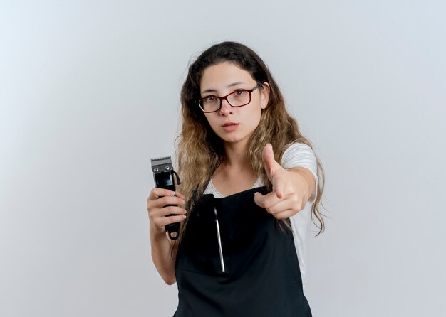 Giovane parrucchiere professionista donna in grembiule tenendo il trimmer che punta con il dito indice davanti guardando fiducioso in piedi sopra il muro bianco