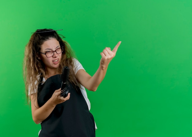 Giovane parrucchiere professionista donna in grembiule tenendo le spazzole per capelli guardando da parte puntando con il dito indice a qualcosa di essere scontento in piedi sopra la parete verde