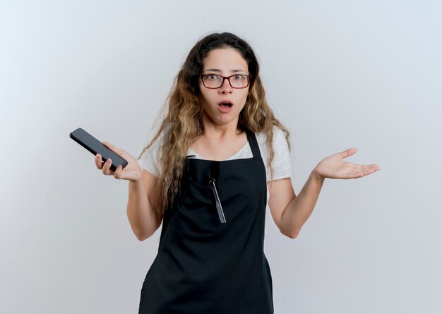 Giovane parrucchiere professionista donna in grembiule tenendo lo smartphone guardando davanti scrollare le spalle essendo confuso e dispiaciuto in piedi sopra il muro bianco