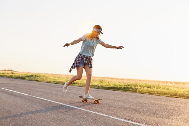 Giovane pattinatrice attraente che guida all'aperto su strada asfaltata, alzò le braccia, donna sportiva che indossa abbigliamento casual che fa skateboard da sola al tramonto in estate.