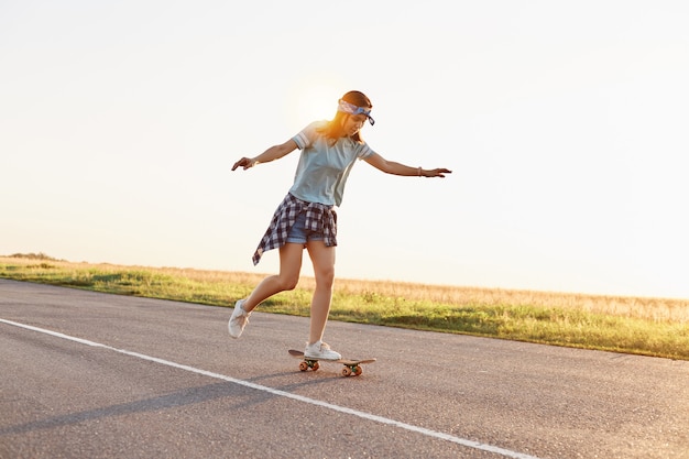 Giovane pattinatrice attraente che guida all'aperto su strada asfaltata, alzò le braccia, donna sportiva che indossa abbigliamento casual che fa skateboard da sola al tramonto in estate.