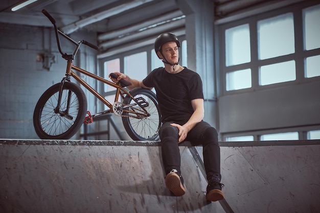 Giovane pilota di Bmx che si rilassa dopo aver praticato trucchi appoggiandosi alla sua bici e distogliendo lo sguardo in uno skatepark al chiuso