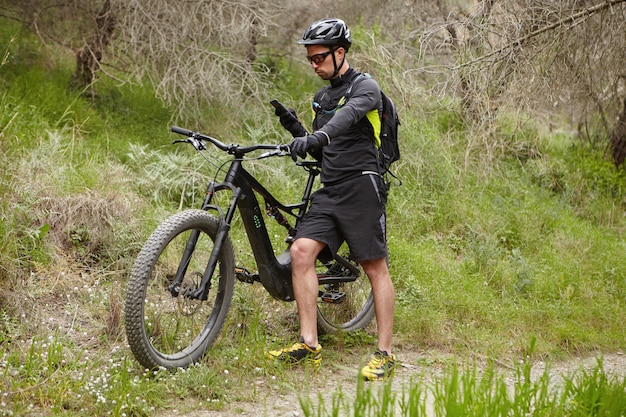 Giovane pilota professionista vestito in abbigliamento da ciclismo e indumenti protettivi alla ricerca di coordinate GPS utilizzando il navigatore sul suo smartphone mentre si guida in bicicletta a batteria nella foresta in una giornata di sole