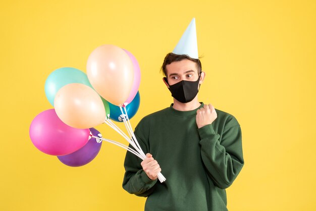 Giovane premuroso di vista frontale con cappuccio del partito e palloncini colorati in piedi sul giallo