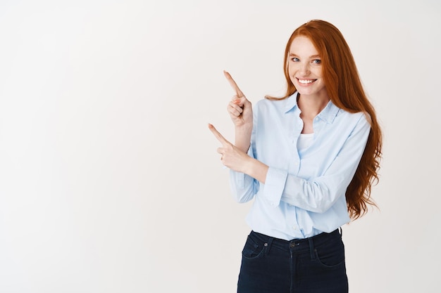 Giovane professionista femminile che indica le dita nell'angolo in alto a sinistra e sorridente. Donna d'affari rossa che mostra il logo sul muro bianco