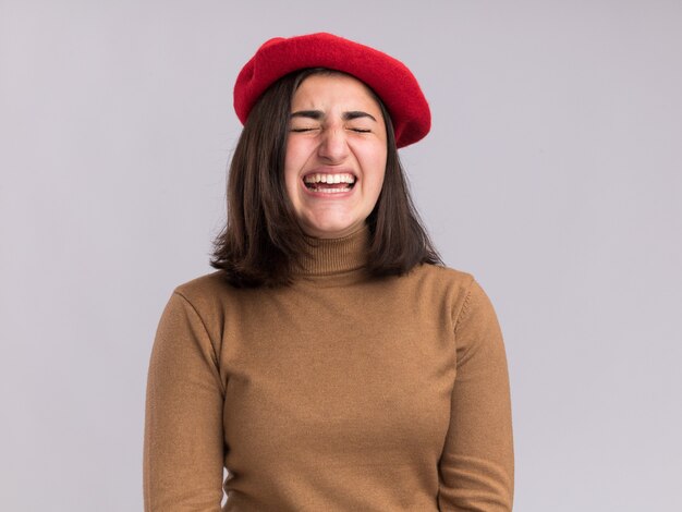 Giovane ragazza abbastanza caucasica scontenta con il cappello del berretto che sta con gli occhi chiusi isolati sulla parete bianca con lo spazio della copia