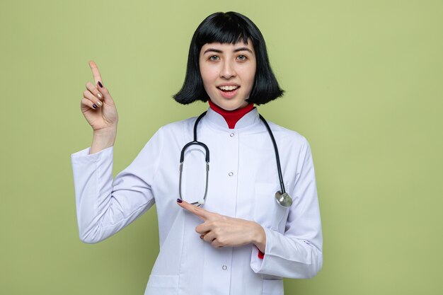 Giovane ragazza abbastanza caucasica sorpresa in uniforme del medico con lo stetoscopio che indica al lato