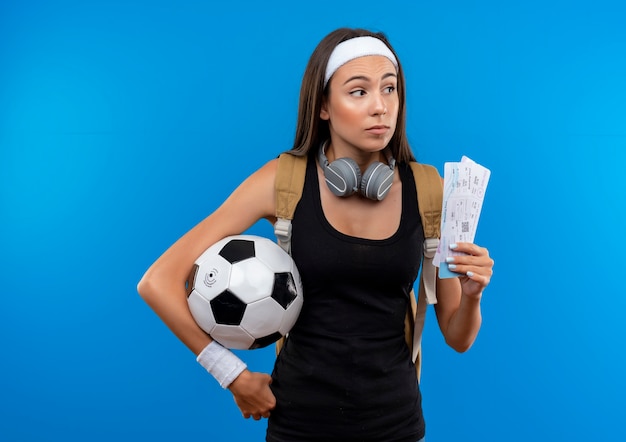 Giovane ragazza abbastanza sportiva che indossa la fascia e il braccialetto e la borsa posteriore con le cuffie sul collo che tiene i biglietti aerei con il pallone da calcio e guardando a lato