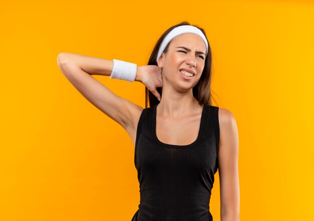 Giovane ragazza abbastanza sportiva dolorante che indossa fascia e cinturino che mette la mano sul collo guardando il lato isolato sul muro arancione