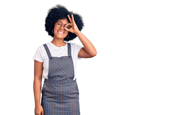 Giovane ragazza afroamericana che indossa abiti casual facendo un gesto ok con la mano sorridente, occhio che guarda attraverso le dita con la faccia felice.