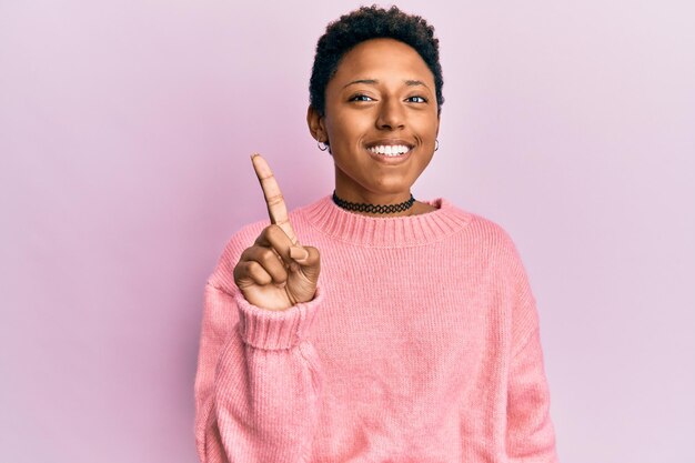 Giovane ragazza afroamericana che indossa abiti casual sorridenti con un'idea o una domanda che punta il dito verso l'alto con la faccia felice, numero uno