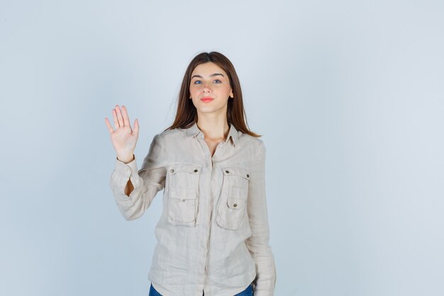 Giovane ragazza agitando la mano per salutare in camicia beige, jeans e dall'aspetto carino. vista frontale.