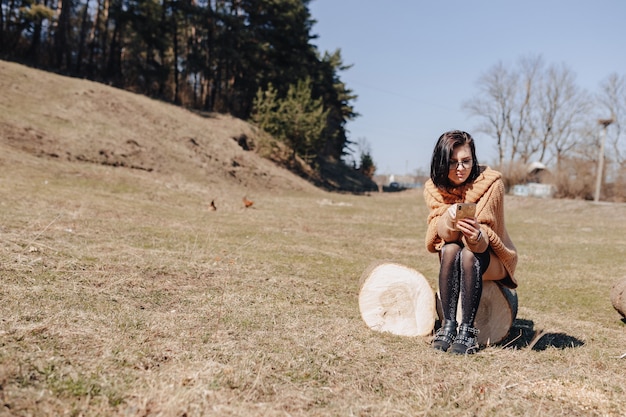 Giovane ragazza alla moda attraente sulla natura sullo sfondo della foresta con il telefono in una giornata di sole