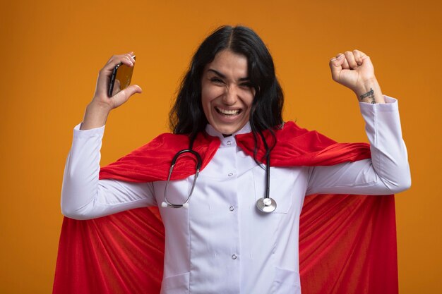 Giovane ragazza allegra del supereroe che indossa la veste medica con lo stetoscopio che tiene il telefono che mostra sì gesto isolato sulla parete arancione