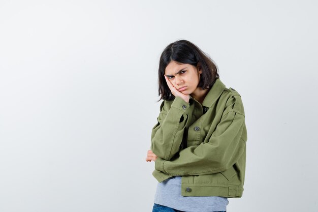 Giovane ragazza appoggiata guancia sul palmo, in piedi in posa di pensiero in maglione grigio, giacca kaki, pantaloni di jeans e guardando pensieroso. vista frontale.