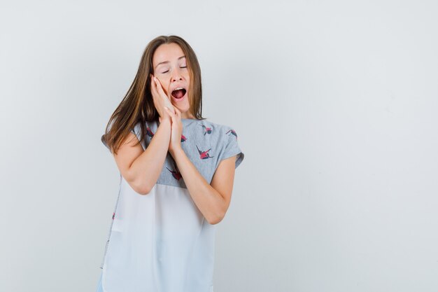 Giovane ragazza appoggiata sul palmo come cuscino e sbadigli in t-shirt vista frontale.