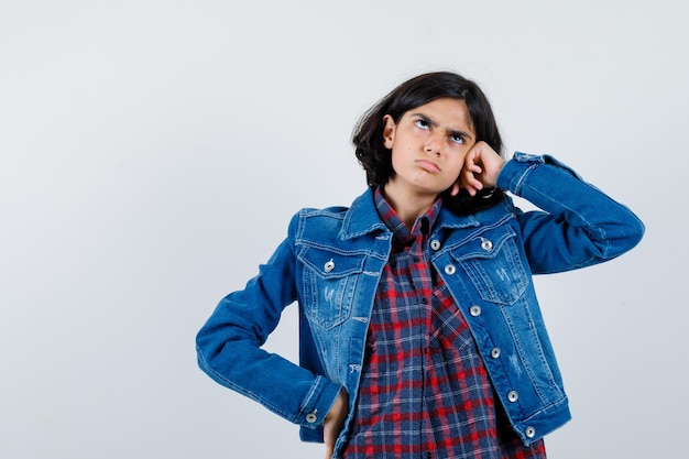 Giovane ragazza appoggiata sulla guancia mentre tiene la mano sulla vita in camicia a quadri e giacca di jeans e sembra pensierosa. vista frontale.