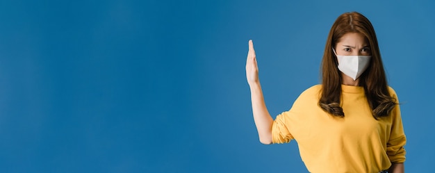 Giovane ragazza Asia indossare la maschera per il viso facendo smettere di cantare con il palmo della mano con espressione negativa e guardando la fotocamera. Allontanamento sociale, quarantena per il virus corona. Banner panoramico sfondo blu.