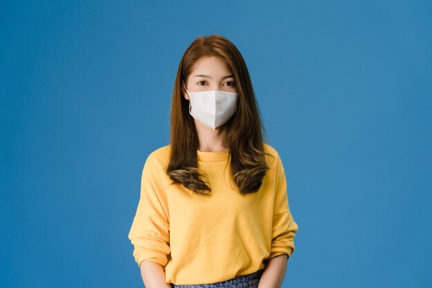 Giovane ragazza asiatica che indossa la maschera medica con vestito in abbigliamento casual e guardando la telecamera isolata su sfondo blu. Autoisolamento, allontanamento sociale, quarantena per la prevenzione del coronavirus.