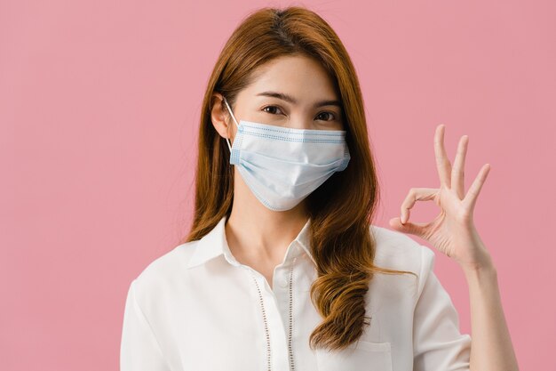 Giovane ragazza asiatica che indossa una maschera medica che gesticola segno ok con un vestito casual e guarda la telecamera isolata su sfondo rosa.