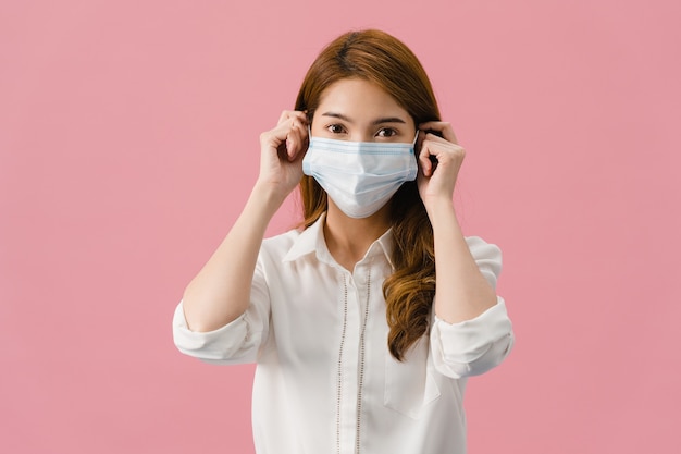 Giovane ragazza asiatica che indossa una maschera medica con abiti casual e guarda la telecamera isolata su sfondo rosa.