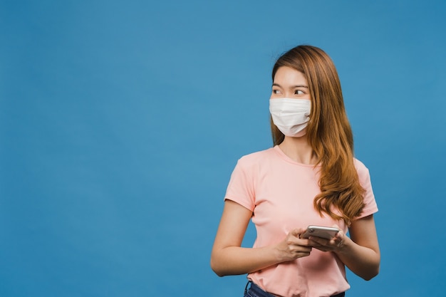 Giovane ragazza asiatica che indossa una maschera per il viso medica usando il telefono cellulare con vestiti in abiti casual isolati sul muro blu