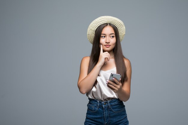 Giovane ragazza asiatica con il telefono di uso del cappello di paglia su fondo grigio