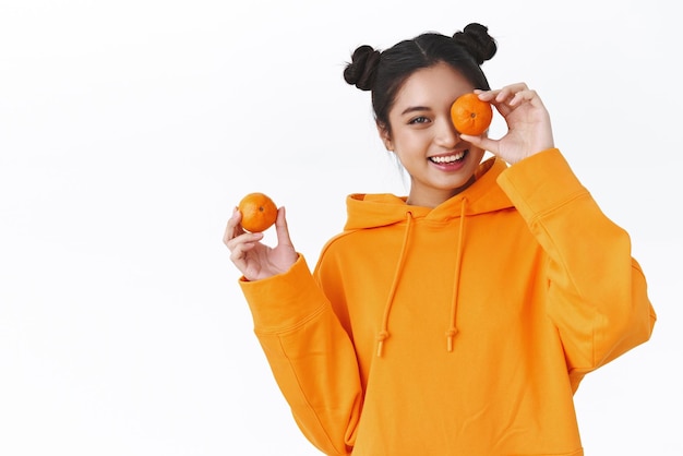 Giovane ragazza asiatica di bell'aspetto spensierata con due ciambelle con cappuccio arancione che tiene i mandarini e sorride giocosamente mangiando frutta prendendosi cura della salute applica cosmetici naturali sfondo bianco