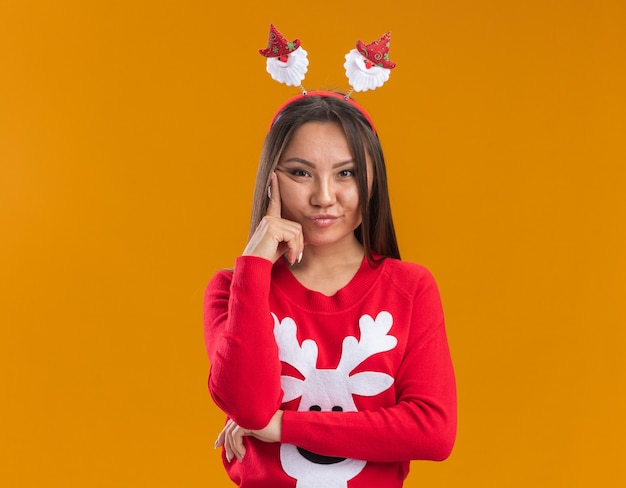 Giovane ragazza asiatica di pensiero che porta il cerchio dei capelli di natale con il maglione che mette il dito sulla guancia isolata sulla parete arancione