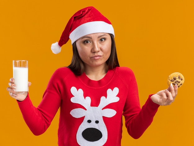 Giovane ragazza asiatica dispiaciuta che porta il cappello di natale con il maglione che tiene un bicchiere di latte con i biscotti isolati sulla parete arancione