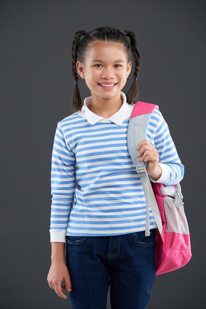Giovane ragazza asiatica in maglione a strisce che posa con lo zaino su una spalla