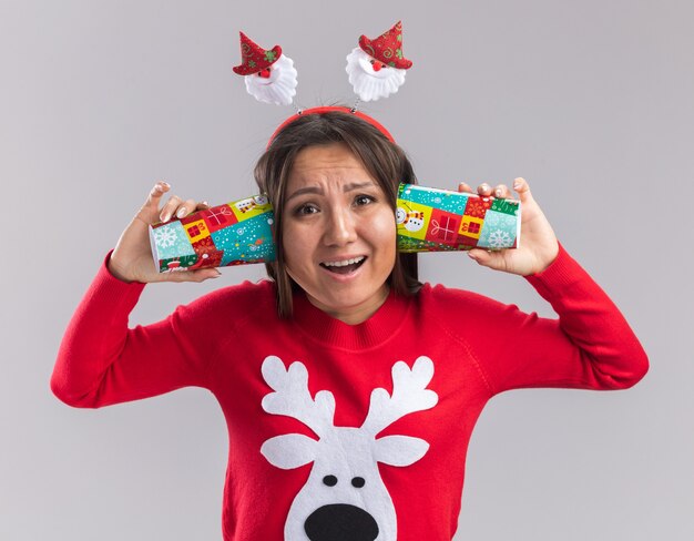 Giovane ragazza asiatica interessata che porta il cerchio dei capelli di natale con il maglione che mette la tazza di natale sulle orecchie isolate su fondo bianco