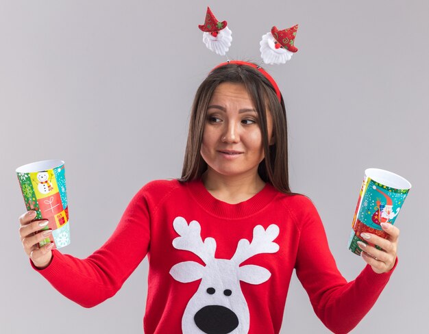 Giovane ragazza asiatica interessata che porta il cerchio dei capelli di natale con la tenuta del maglione e l'esame della tazza di natale isolata su fondo bianco