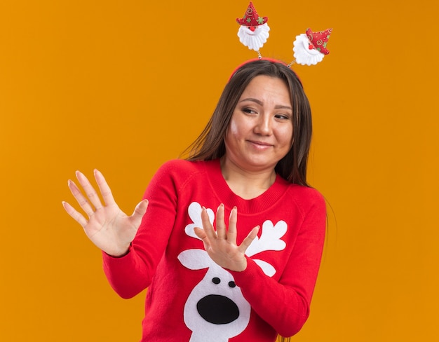 Giovane ragazza asiatica schizzinosa che indossa il cerchio dei capelli di Natale con il maglione che tiene le mani sul lato isolato sulla parete arancione