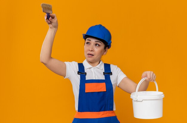 Giovane ragazza asiatica scontenta del costruttore con il casco di sicurezza blu che tiene il pennello e la pittura ad olio