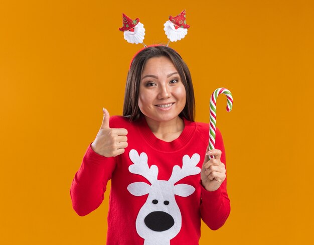 Giovane ragazza asiatica sorridente che porta il cerchio dei capelli di natale con la caramella di natale della tenuta del maglione che mostra pollice su isolato sulla parete arancione