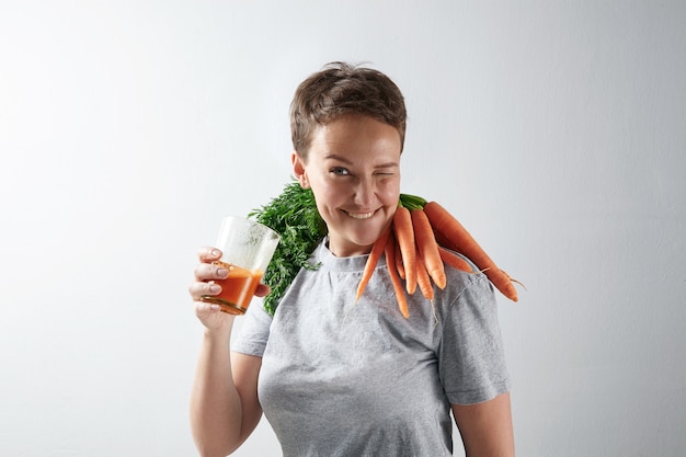 Giovane ragazza attraente con una pelle sana e perfetta sbatte le palpebre felicemente mentre beve il suo succo di carota biologico fresco con il raccolto di carote sulle spalle