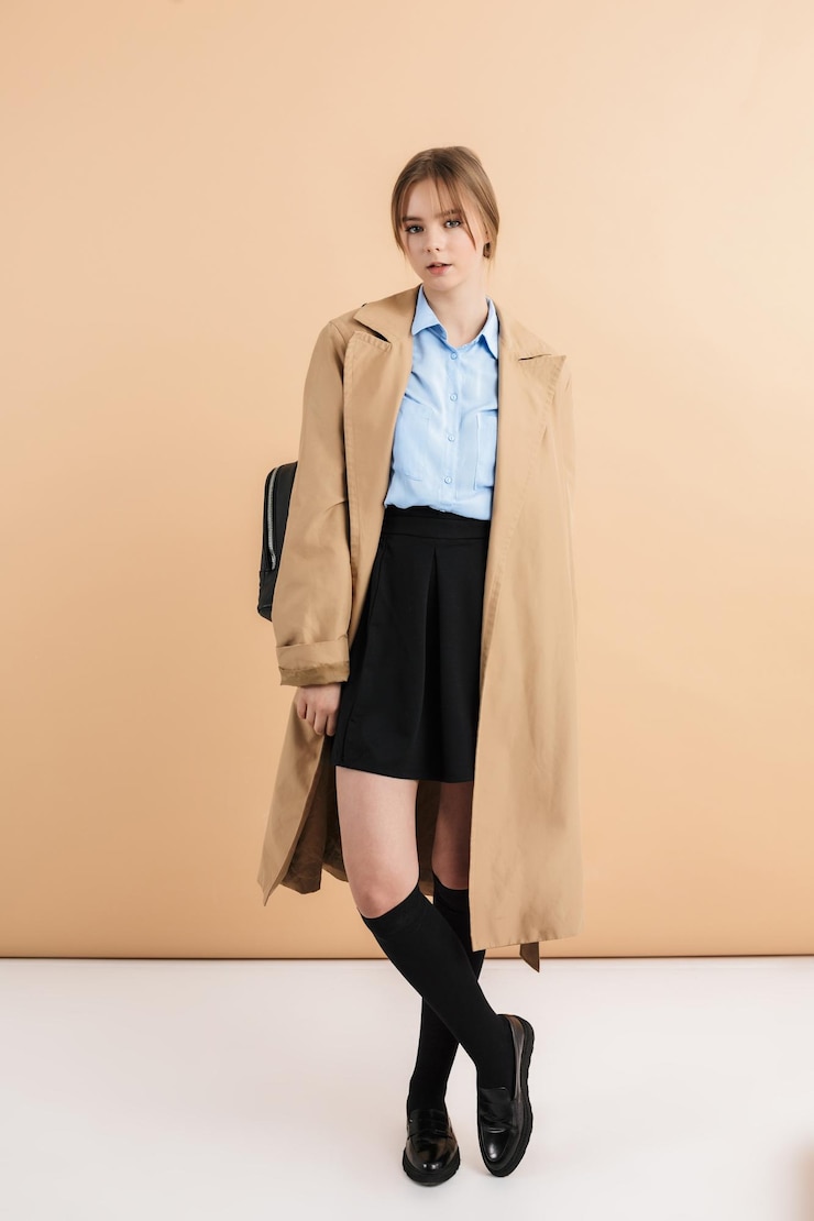 Foto gratuito giovane ragazza attraente in trench e camicia blu con zaino sulla spalla che guarda sognante a porte chiuse su sfondo beige isolato
