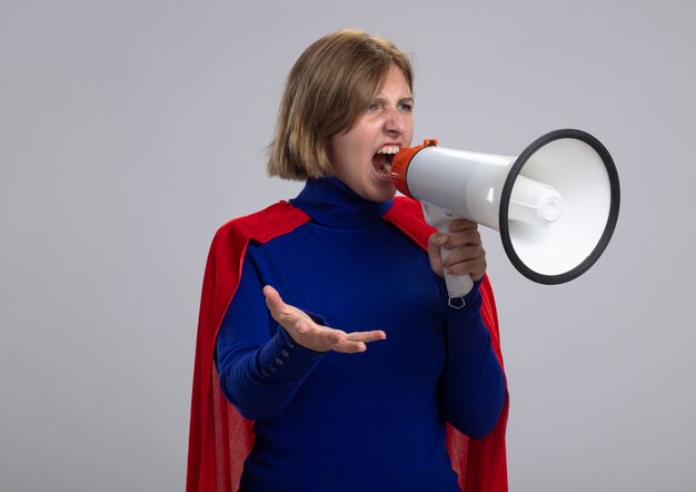 Giovane ragazza bionda arrabbiata del supereroe in mantello rosso che grida in altoparlante guardando dritto mostrando la mano vuota isolata sul muro bianco con lo spazio della copia