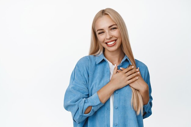 Giovane ragazza bionda che sembra lusingata tenendosi per mano sul cuore e sorridente essendo toccata o affezionata a qualcosa in piedi su sfondo bianco