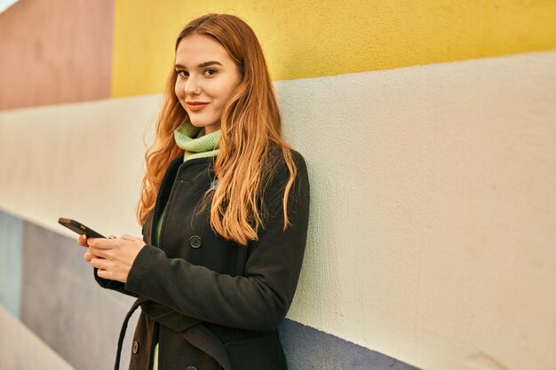 Giovane ragazza bionda che sorride felice utilizzando lo smartphone in città