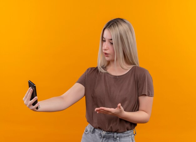 Giovane ragazza bionda che tiene il telefono cellulare alla ricerca e indicando con la mano sulla parete arancione isolata