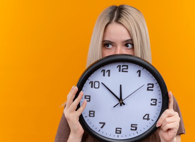 Giovane ragazza bionda che tiene l'orologio che si nasconde dietro di esso guardando il lato sinistro sulla parete arancione isolata con lo spazio della copia