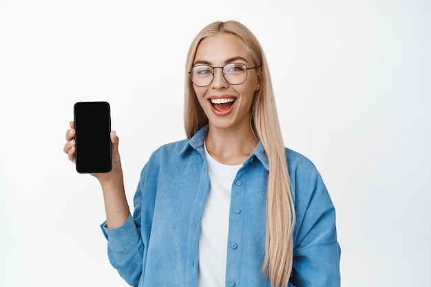 Giovane ragazza bionda con gli occhiali che mostra lo schermo del telefono cellulare e sorridente, in piedi in abiti casual, dimostrando l'applicazione per smartphone su bianco.