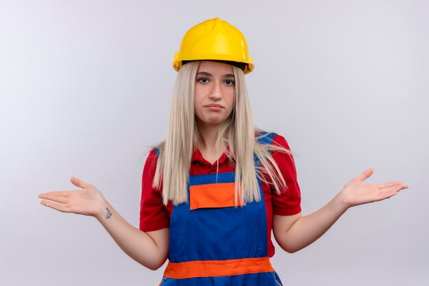 Giovane ragazza bionda confusa del costruttore dell'ingegnere in uniforme che mostra le mani vuote sullo spazio bianco isolato