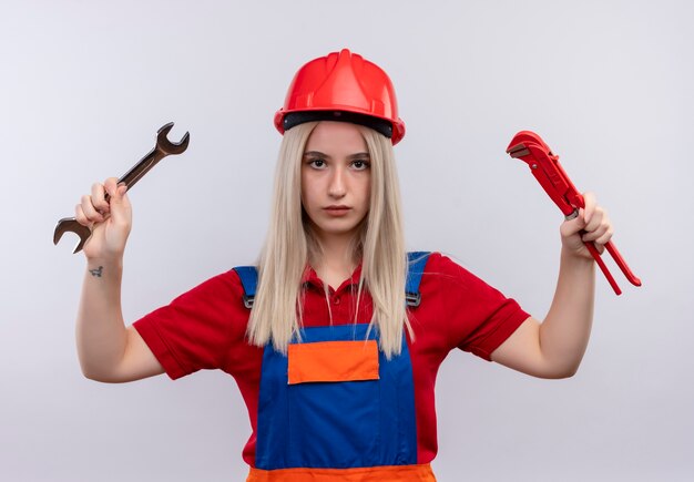 Giovane ragazza bionda del costruttore dell'ingegnere in uniforme che tiene la chiave aperta e la chiave a tubo che osserva sulla parete bianca isolata